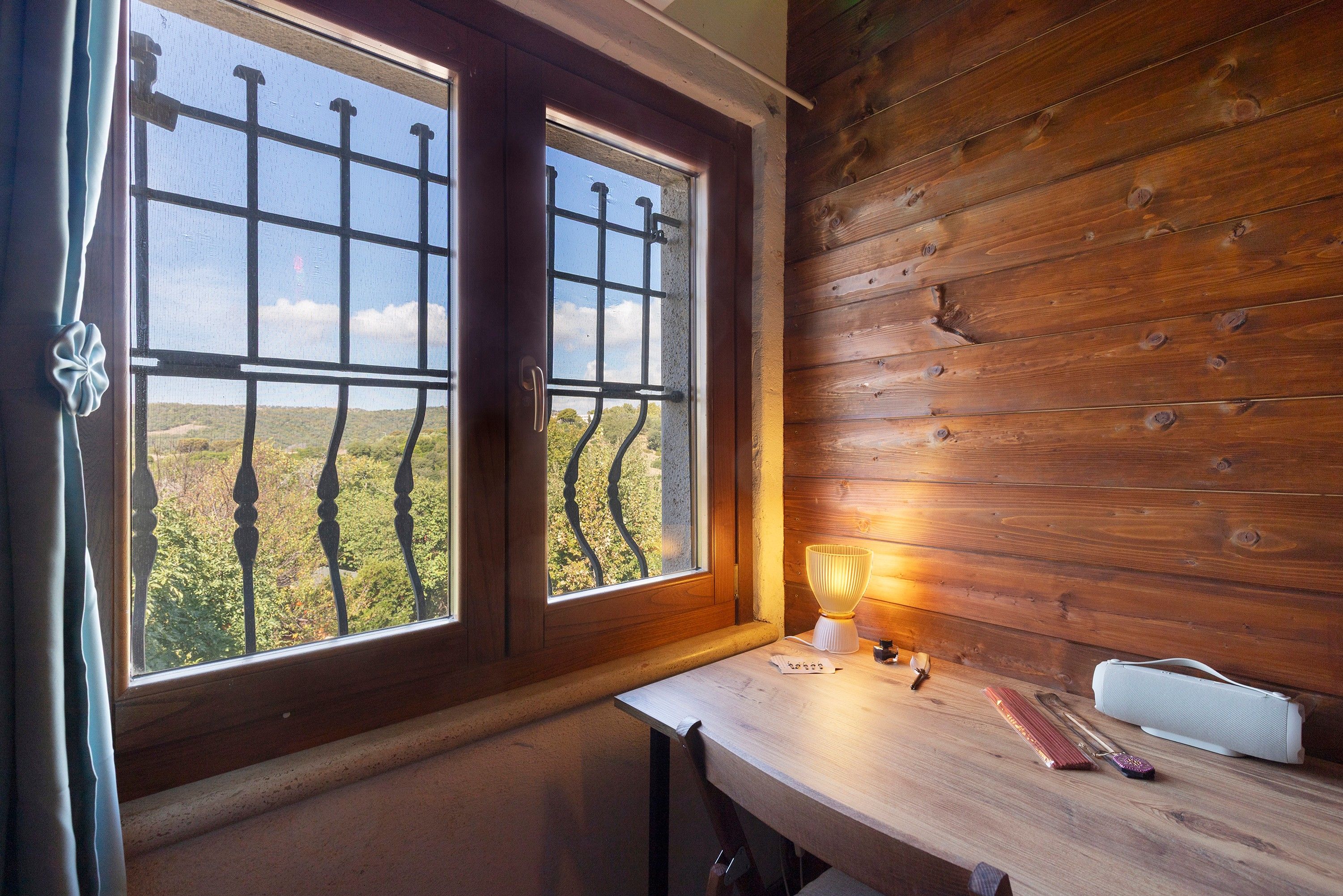 Ein Zimmer mit zwei Fenstern, einem Holztisch mit Lampe und Blick auf eine Landschaft.