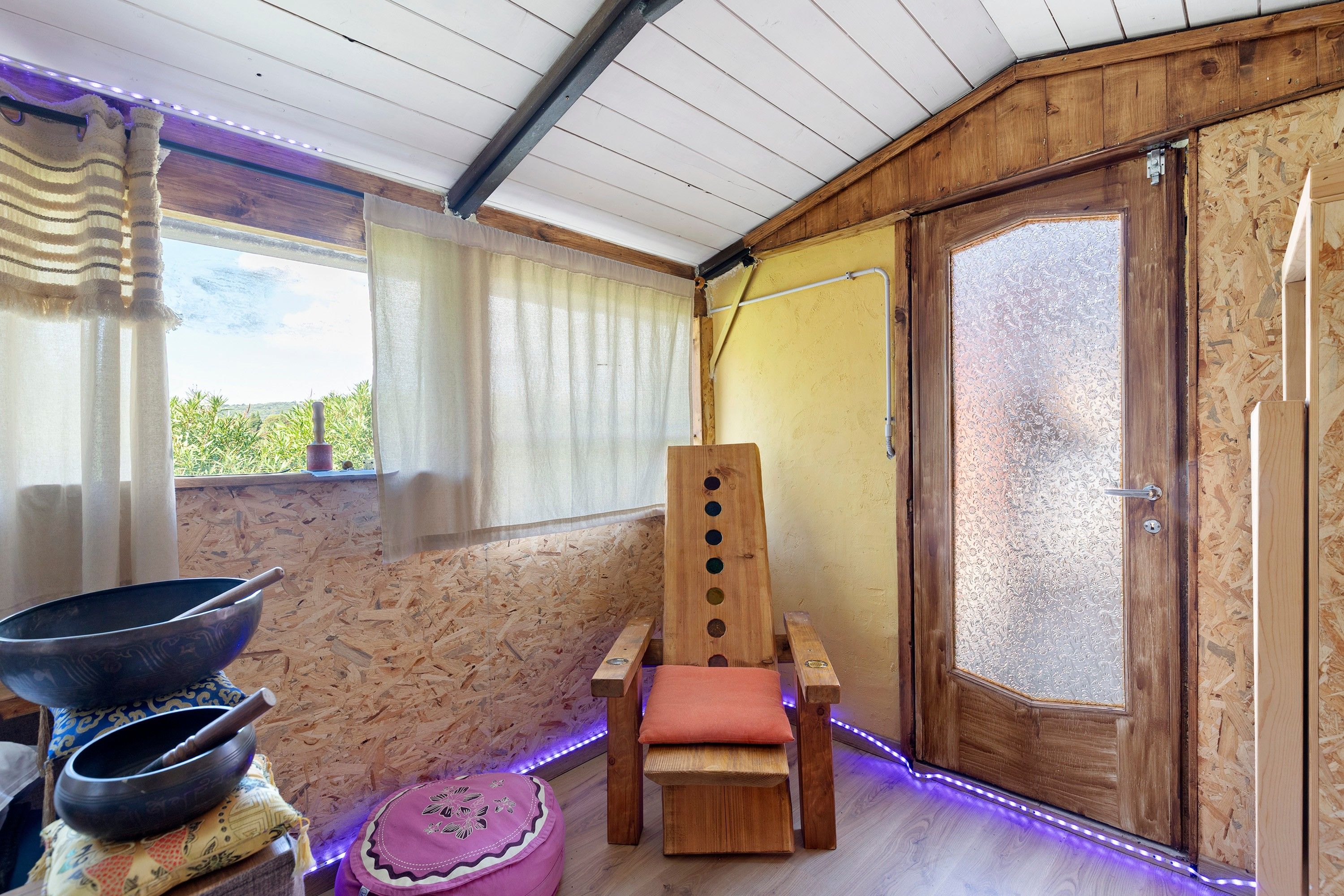 Ein Zimmer mit einem Holzstuhl, einem Vorhangfenster, einem Holzboden und Blick auf Grünfläche.