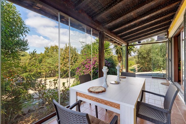 A covered outdoor dining area with a table, chairs, and a view of the surrounding garden.