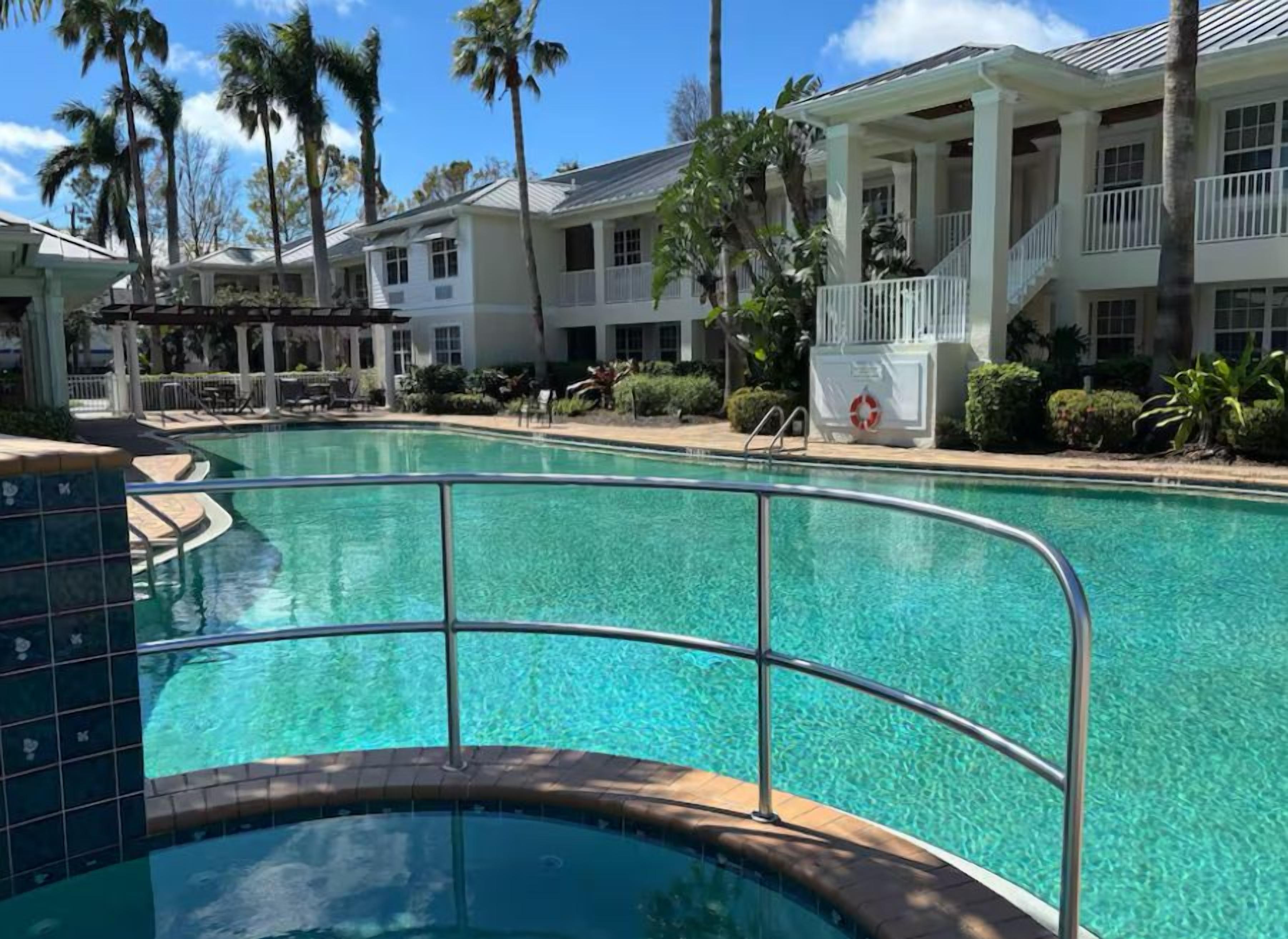 A swimming pool with a tiled deck, surrounded by a building with balconies and palm trees.