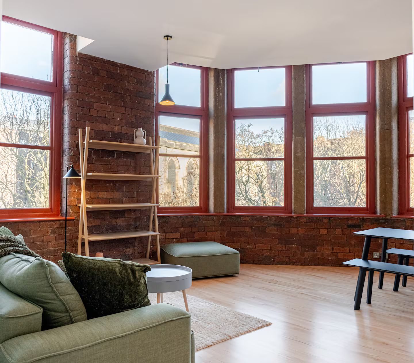 A room with a brick wall, large windows, a sofa, a table, and a lamp.
