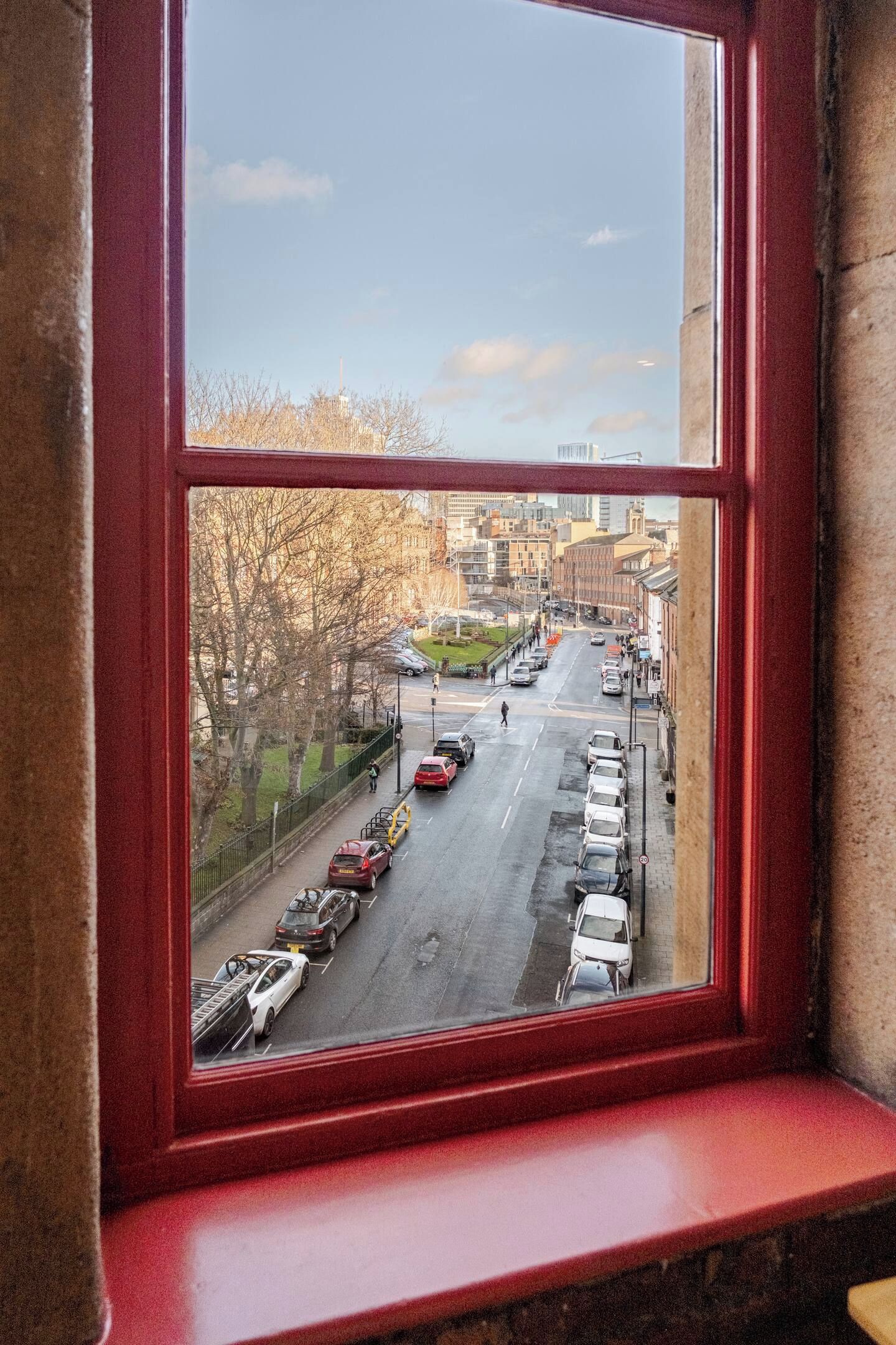 A view through a window with a red frame, looking out onto a city street with parked cars and buildings.