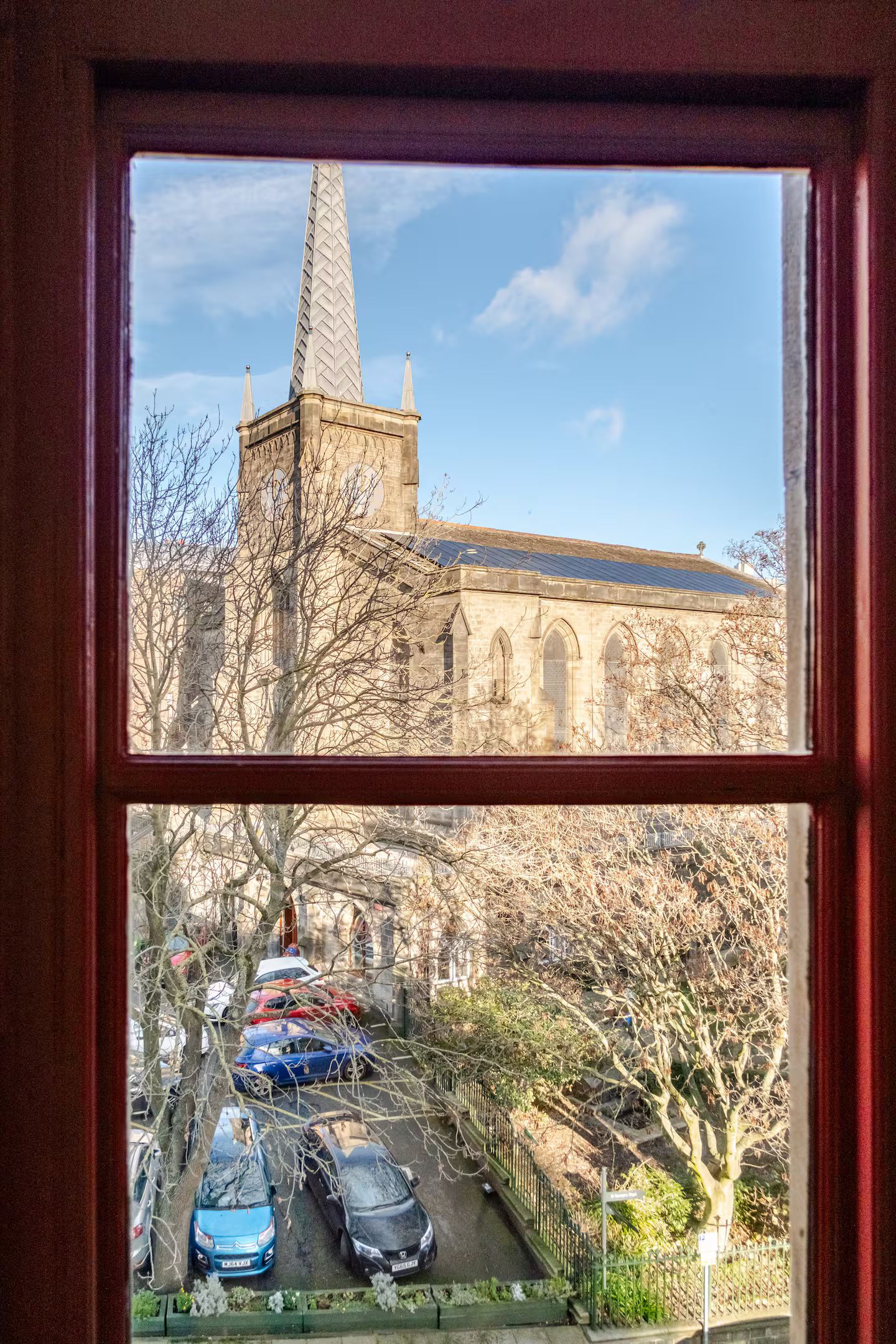 A view through a window of a building with a tall spire, parked cars, and leafless trees.