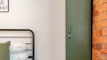 Un dormitorio con una cama, un taburete pequeño, un casillero de metal verde y una pared de ladrillo.