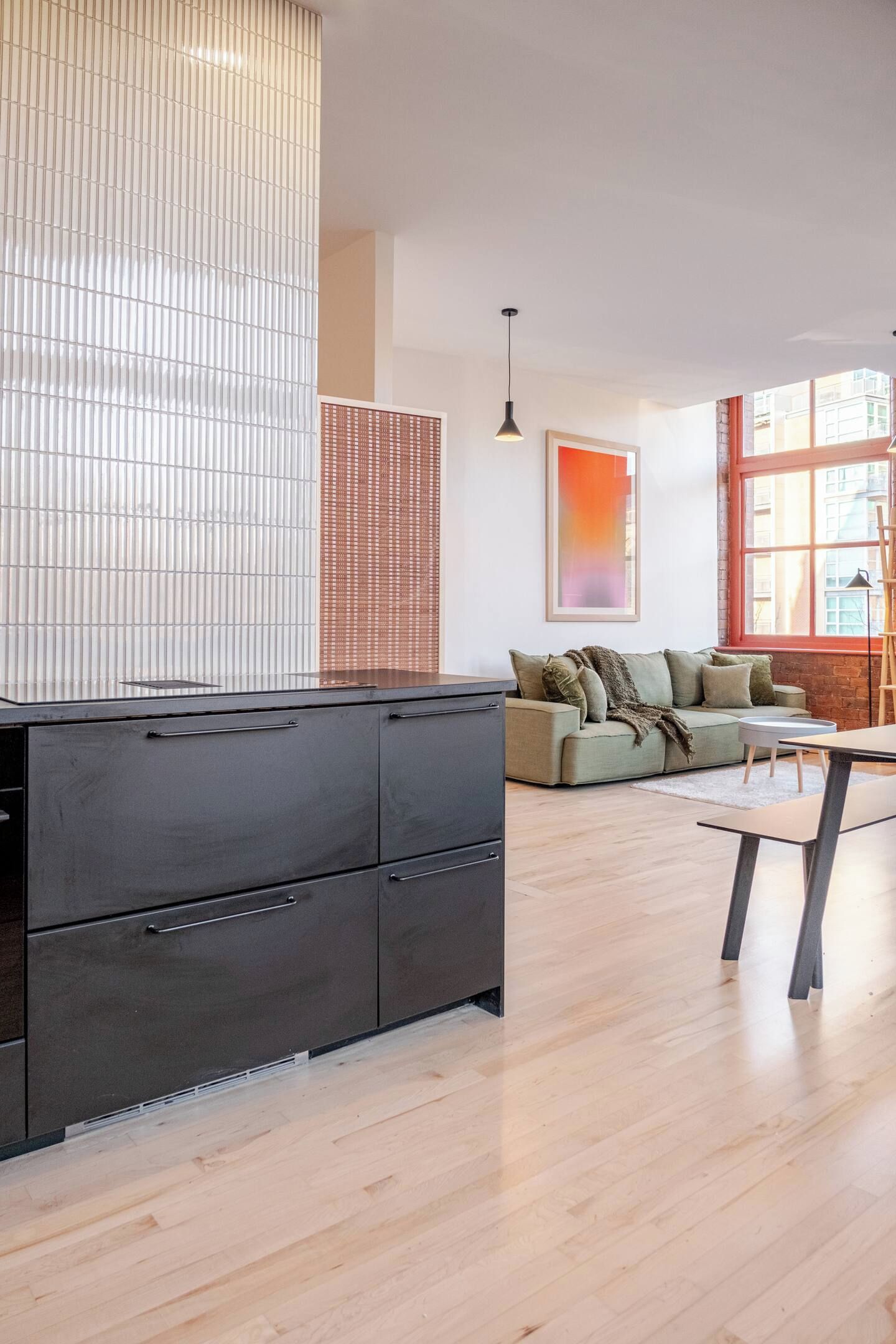 A modern kitchen with a black countertop and cabinets, a wooden floor, a sofa with cushions, and a large window with a view.