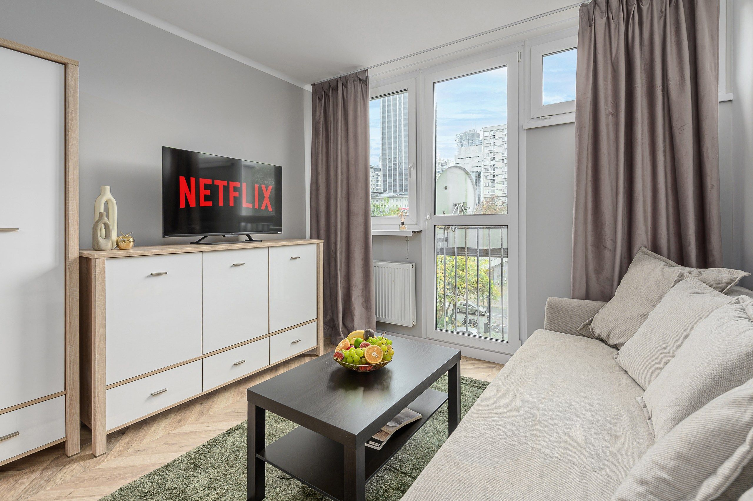A modern living room with a flat-screen TV displaying the Netflix logo, a sofa, a coffee table with a fruit bowl, and a view of the city through large windows.