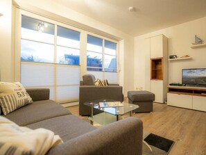 A modern living room with a sofa, armchair, and ottoman, a glass coffee table, a flat-screen TV, and a large window with blinds. - 6 in the Pole Poppenspaler House (St. Peter-Ording)