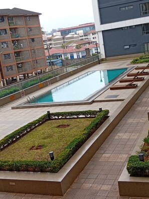 Ein Pool mit Liegestühlen, ein angelegter Gartenbereich und moderne Gebäude im Hintergrund.