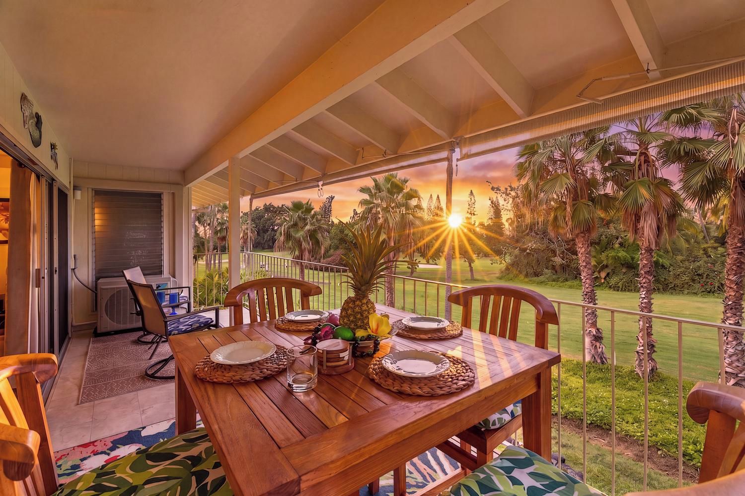 A wooden dining table set for two with a tropical fruit centerpiece, overlooking a lush garden and a sunset view.