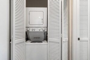 A compact, enclosed laundry room with a washing machine and dryer, featuring a small window and control knobs. - Keauhou Palena #604 – Peaceful 1 Bedroom, 2 Bath Kona Retreat with A/C (KAILUA-KONA)