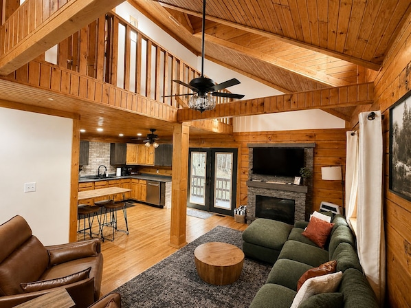A spacious living area with a high ceiling, wooden beams, a fireplace, a television, and a green sofa.