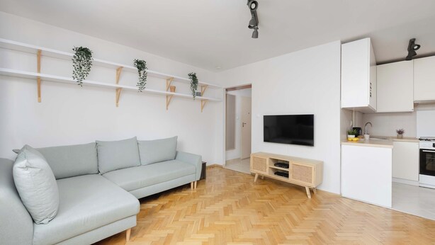 Ein modernes Wohnzimmer mit grauem Sofa, Holzboden, weißen Wänden, einem Fernseher und einer Küchenzeile mit Schränken und Herd.