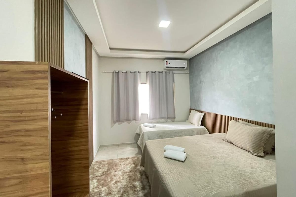A hotel room with two beds, a wardrobe, an air conditioning unit, and a window with curtains.