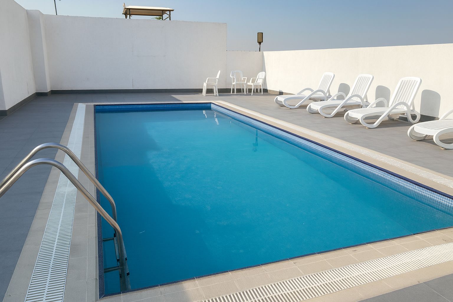 Una piscina rectangular con una escalera metálica a un lado, rodeada de sillones blancos y un muro blanco.