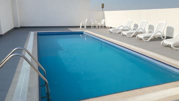 Una piscina rectangular con una escalera metálica a un lado, rodeada de sillones blancos y un muro blanco.
