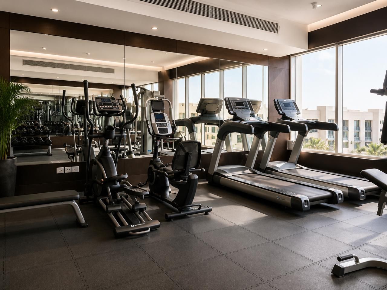 Ett modernt gym med olika typer av träningsutrustning, stora fönster och utsikt över byggnader.