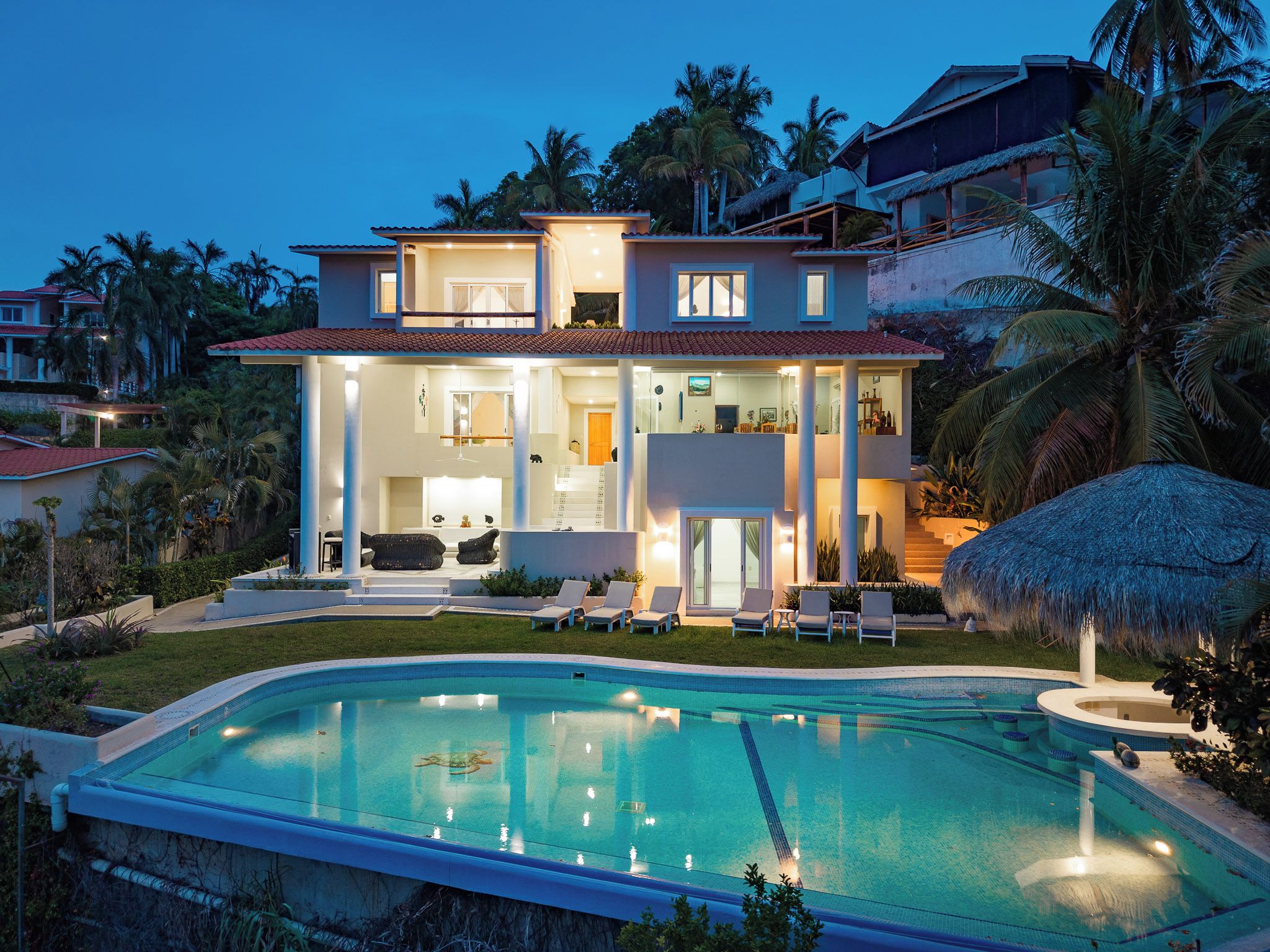 Ein zweistöckiges Haus mit Pool, Sitzgelegenheit im Freien und einem Reetdachschirm.