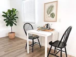 A dining area with a white table, two black chairs, a potted plant, and a framed botanical illustration on the wall.