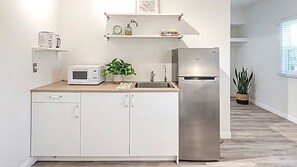 A compact kitchen with white cabinetry, a stainless steel refrigerator, a microwave, and a sink. There are shelves above the counter holding books and a clock.