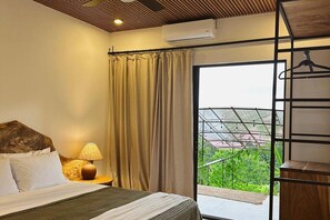 Quarto de hotel com cama, mesa de cabeceira, abajur, janela com cortinas e vista para o verde lá fora.