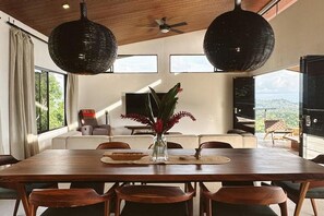 Ein Essbereich mit Holztisch, Stühlen und einer Vase mit Blumen. Im Hintergrund befindet sich eine Couch und ein Fernseher.
