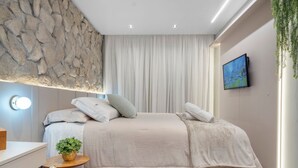 A modern bedroom with a stone wall, a bed with multiple pillows, a bedside table with a plant, a TV on the wall, and a window with curtains. - Apto YOU 206 (Canela)