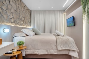 A modern bedroom with a stone wall, a bed with multiple pillows, a bedside table with a plant, a TV on the wall, and a window with curtains. - Apto YOU 206 (Canela)