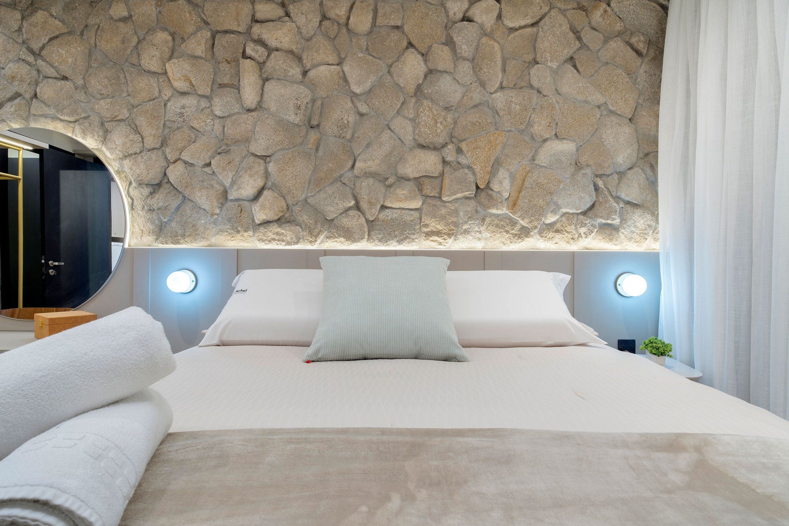 Een slaapkamer met een stenen muur, een bed met witte beddengoed, twee nachtlampjes en een ronde spiegel.