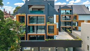 A modern multi-story building with large windows, balconies with greenery, and a visible commercial space on the ground floor. - Apto YOU 206 (Canela)