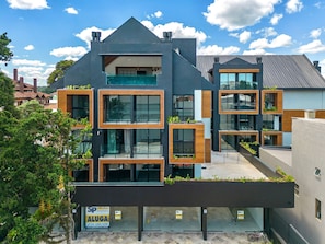 A modern multi-story building with large windows, balconies with greenery, and a visible commercial space on the ground floor. - Apto YOU 206 (Canela)