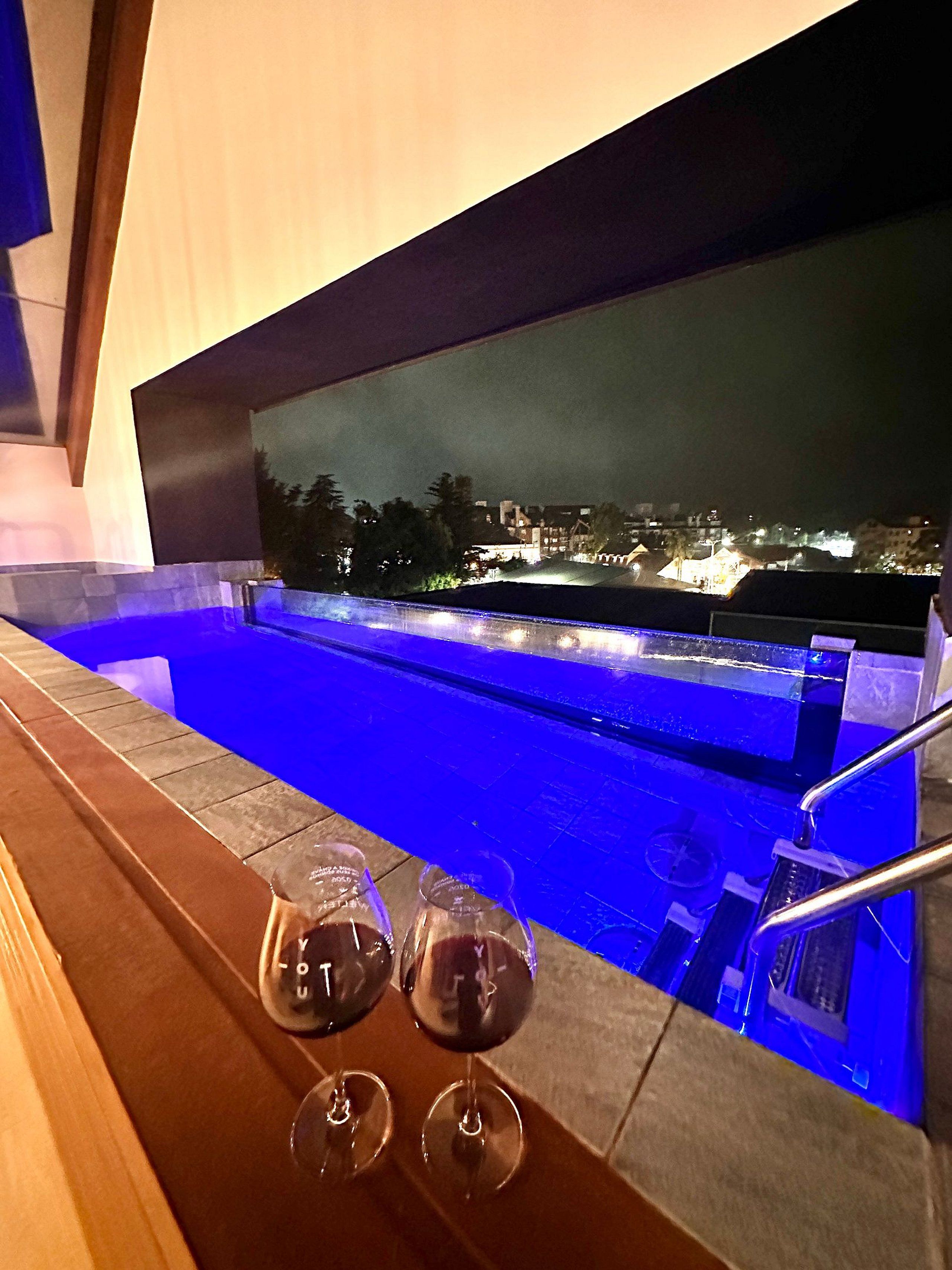 Zona de piscina con vista a un paisaje urbano nocturno, dos copas de vino tinto y un banco de madera.
