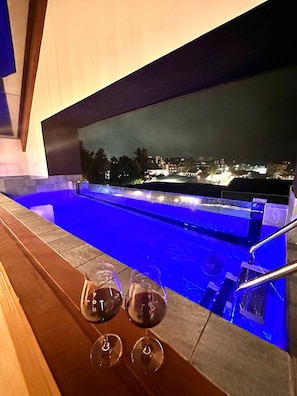 A pool area with a view of a cityscape at night, two glasses of red wine, and a wooden bench. - Apto YOU 206 (Canela)