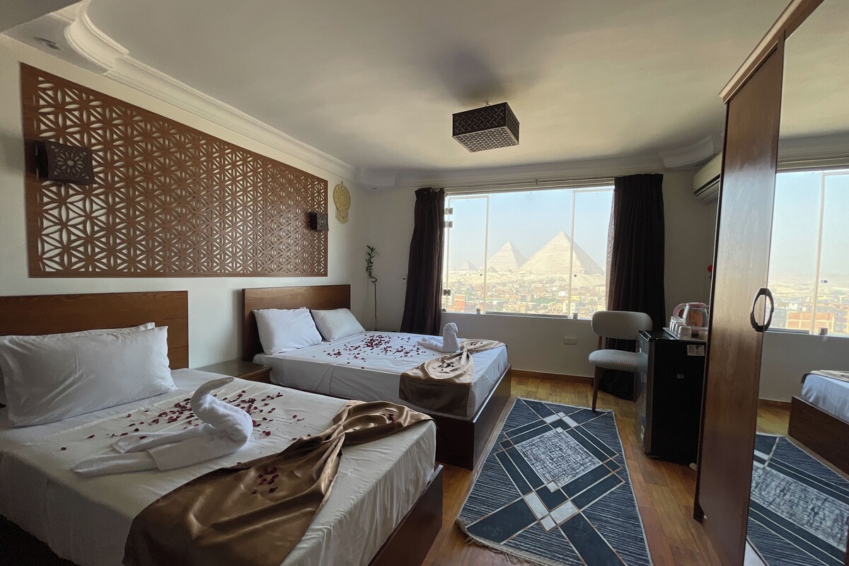 A hotel room with two beds, a view of the pyramids, a decorative wall panel, and a modern ceiling light.