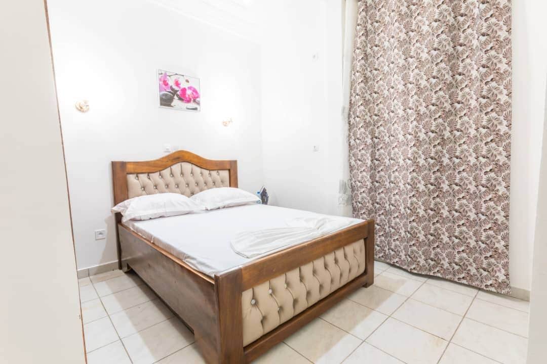 Quarto com cama de madeira, roupa de cama branca, cortina estampada e uma pequena moldura na parede.