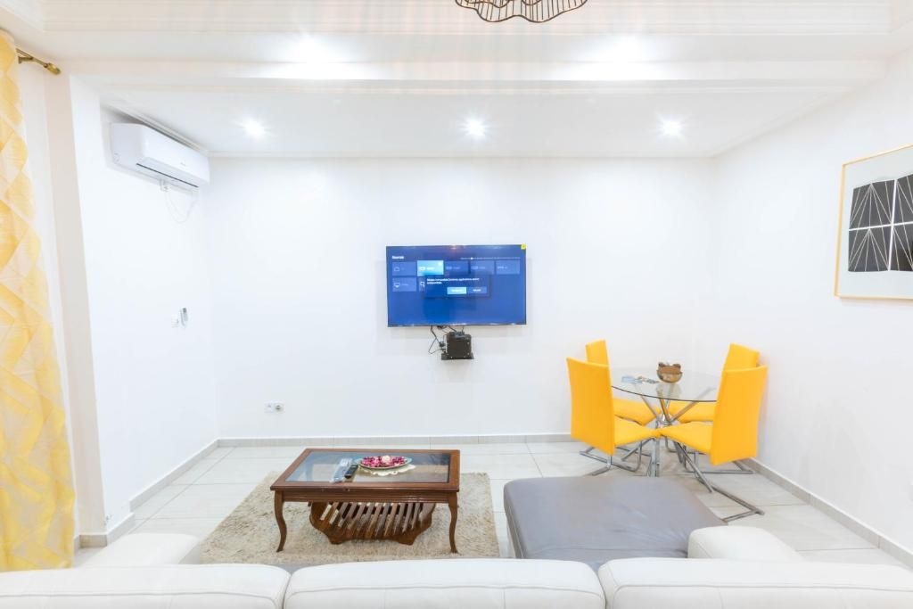 A modern living room with a flat-screen TV mounted on the wall, a coffee table with a bowl and remote, and a seating area with a sofa and yellow chairs.
