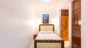 A bedroom with a bed, a nightstand, a poster on the wall, and a wooden door. - EDEN GARDEN LUXURIOUS GUEST HOUSE (Yaoundé)