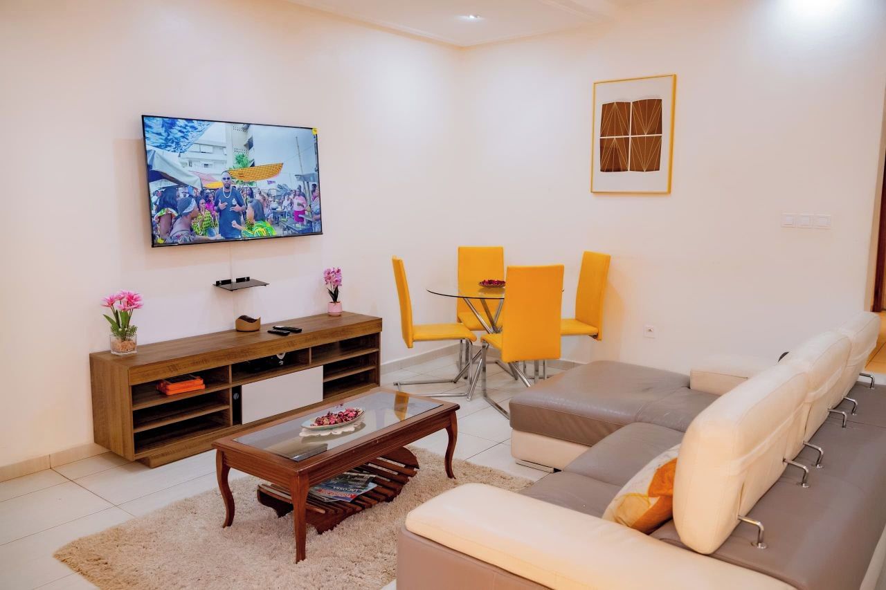 Una sala de estar con un televisor de pantalla plana, un mueble de madera para entretenimiento, un sofá, una mesa de centro y un comedor con sillas amarillas.