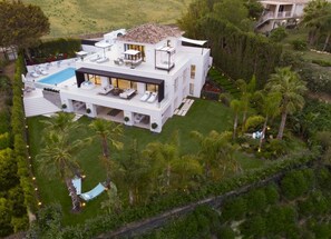 Ein modernes, zweistöckiges Haus mit Pool auf dem Dach, umgeben von üppiger Vegetation und Palmen.