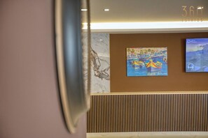Un área de recepción con un panel de madera en la pared, una obra de arte enmarcada que representa barcos y una pantalla de televisión mostrando un paisaje.