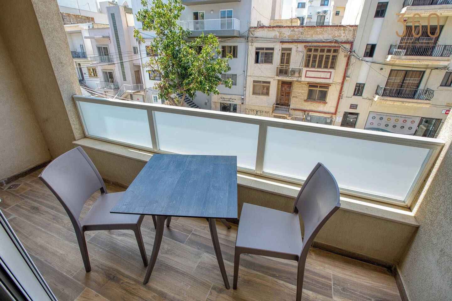 Een balkon met een tafel en twee stoelen, met uitzicht op een stadsgezicht met gebouwen en een boom.