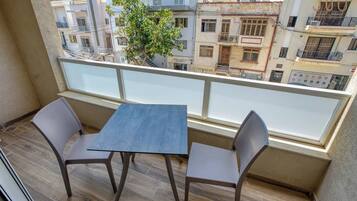 Een balkon met een tafel en twee stoelen, met uitzicht op een stadsgezicht met gebouwen en een boom.