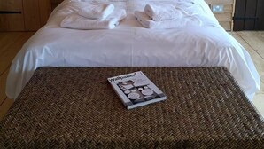 Een net opgemaakt bed met witte lakens en handdoeken, een tijdschrift op een geweven onderzetter, en houten wandbekleding.