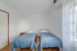 Quarto com duas camas de solteiro, piso de madeira, porta branca e janela com cortinas finas.