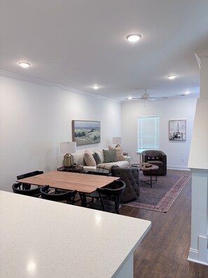 A modern living room with a dining area, white walls, ceiling lights, a large window with blinds, and a ceiling fan. - 2025 Townhome-Style Condo 3 Miles from the Square (Oxford)