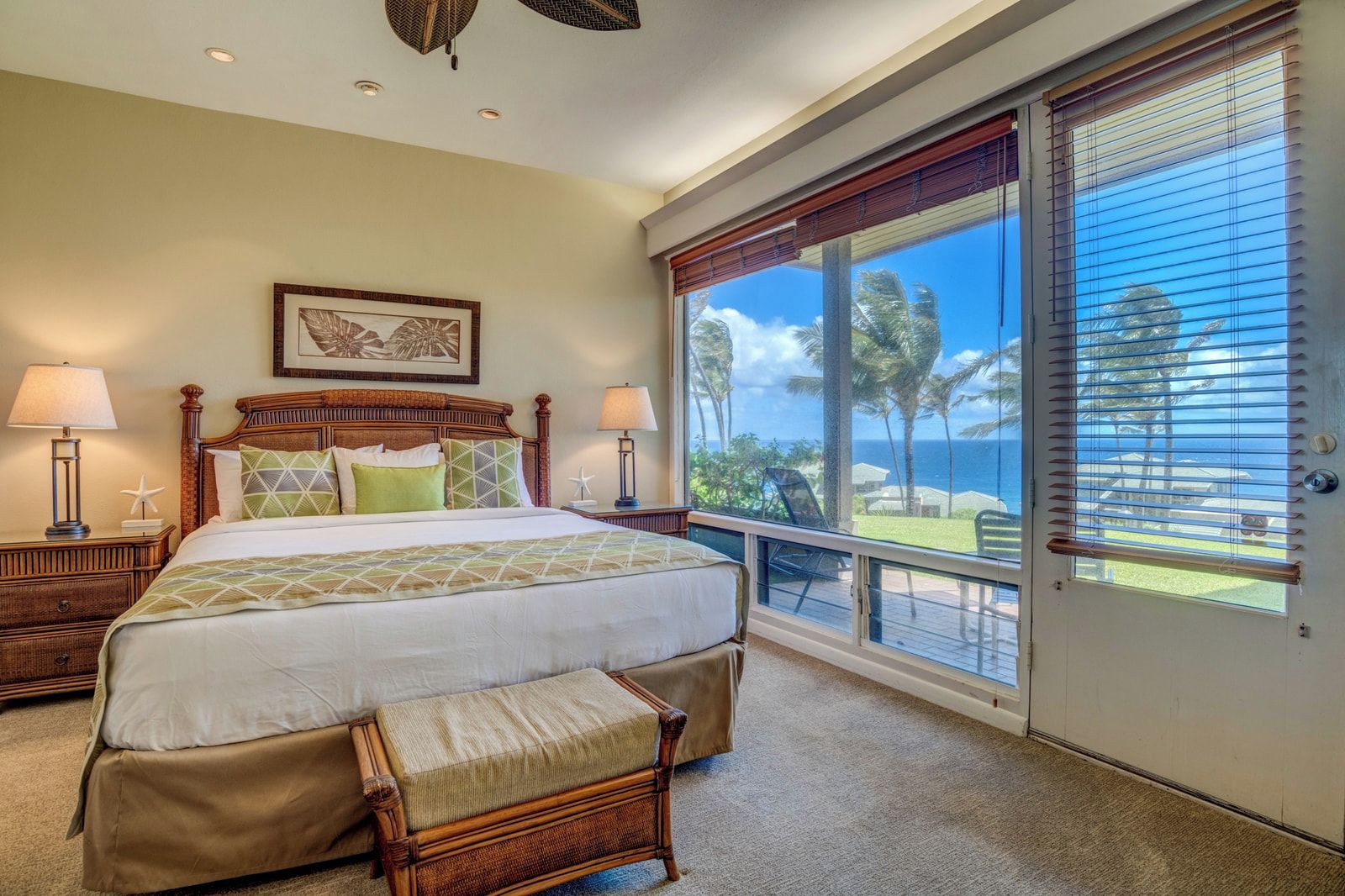 A bedroom with a large bed, two bedside tables, a nightstand, a window with a view of palm trees and the ocean, and a wooden bench.