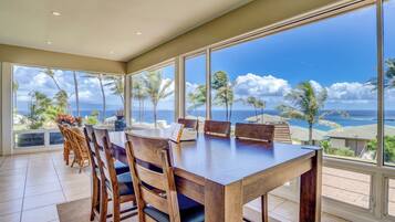 A dining area with a large table and chairs, large windows with a view of the ocean and palm trees.
