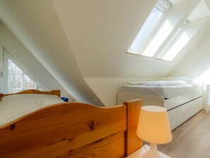 Ein Schlafzimmer mit schrägem Dach, einem Holzbett, einem Nachttisch mit Lampe und einem Fenster, das für natürliches Licht sorgt.