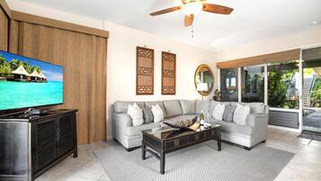 A living room with a grey sofa, a coffee table, a TV, decorative panels, a ceiling fan, and a large window.