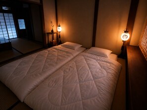 A traditional Japanese-style room with tatami flooring, a large bed with a quilted cover, and two wall-mounted lanterns providing soft lighting.