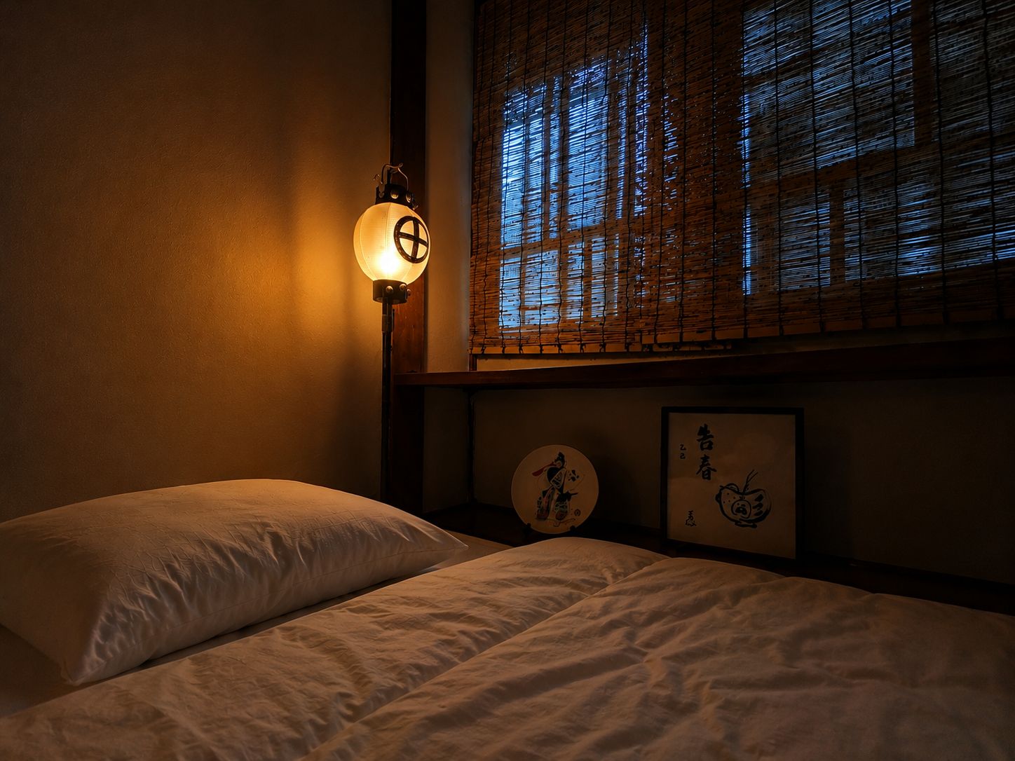 A dimly lit room with a bed, a wall-mounted lamp, and a window with bamboo blinds.